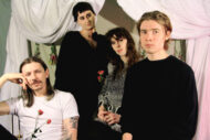 The doom metal band SEED are grouped standing and sitting together. Drummer Chelsea Ellsworth and bassist Jack Whelan are holding roses.
