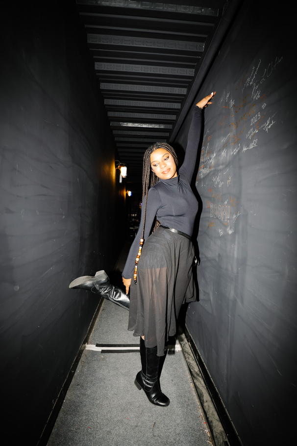 a woman poses in a hallway with her right arm up against the wall and right leg lifted up while no teeth smiling